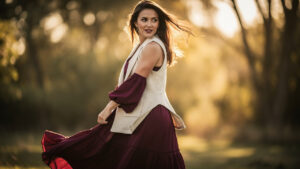 Une femme porte un gilet sans manches crème sur une robe bohème fluide bordeaux, éclairée par la lumière naturelle, avec le tissu légèrement soulevé par le vent.
